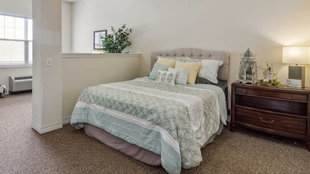A neatly made bed with green and white bedding is next to a wooden nightstand with a lamp and decorative items in a cozy, carpeted bedroom with neutral walls and a window in the background.
