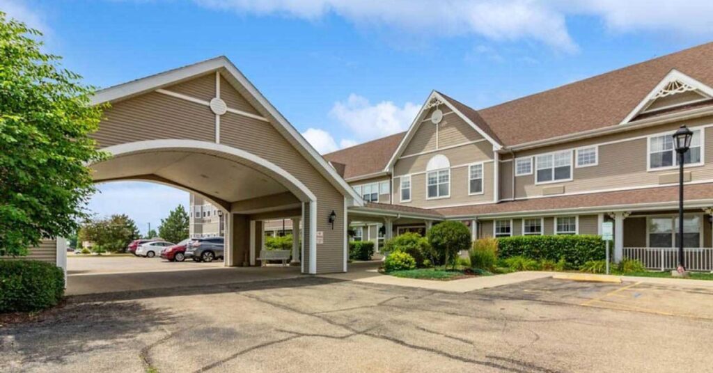 A large, two-story residential building with beige siding, white trim, and a covered driveway entrance. Several cars are parked nearby, with shrubs and greenery lining the walkway. The sky is blue with scattered clouds.