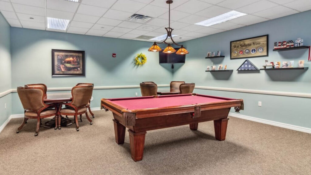 A room with a red pool table in the center, four brown chairs around a round table to the left, and wall shelves holding framed photos, a folded flag, and cards. The walls are light blue and decorated with artwork.