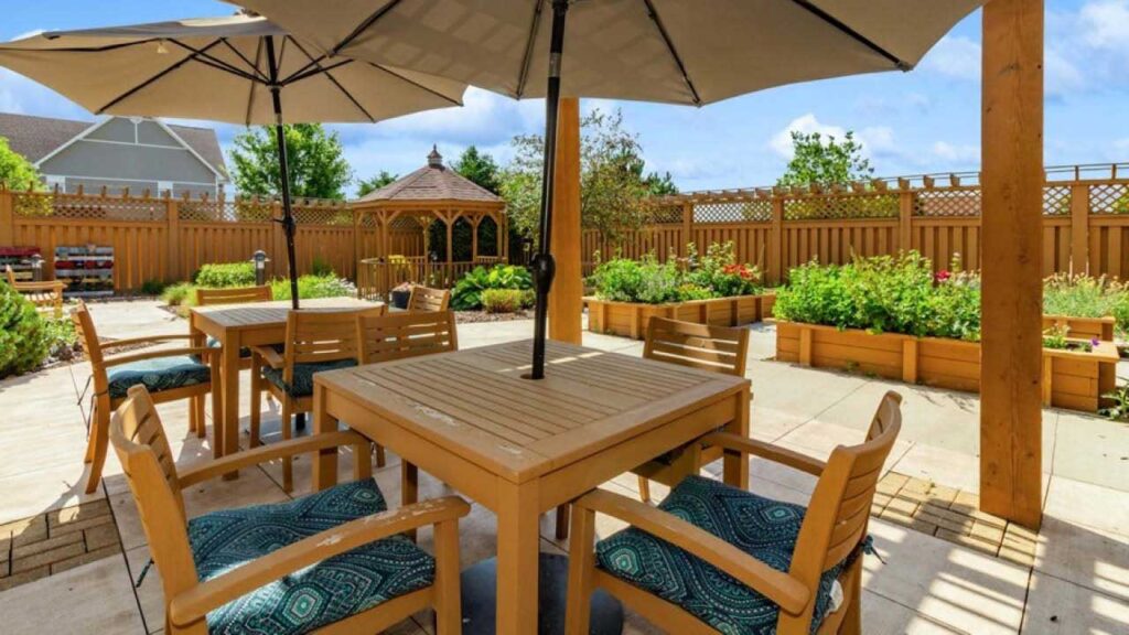 A sunny outdoor patio with wooden tables and chairs under large umbrellas, surrounded by raised garden beds, a wooden fence, and a gazebo in the background. Cushioned seats add comfort to the seating area.