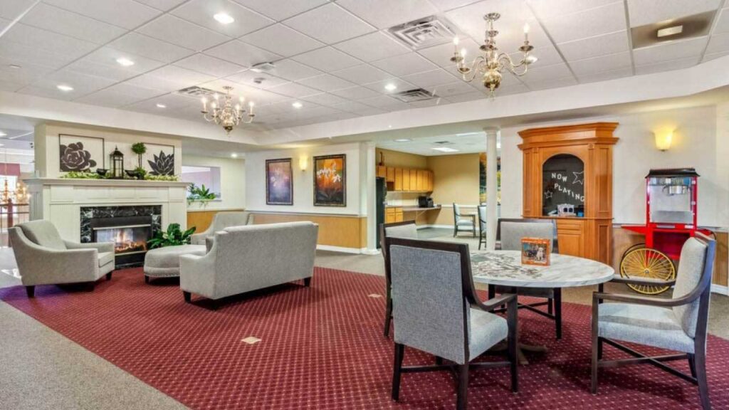 A cozy, well-lit lounge with gray armchairs, a small round table, a fireplace, artwork on the walls, and a chandelier; nearby are a kitchen area and a popcorn machine on a red cart.