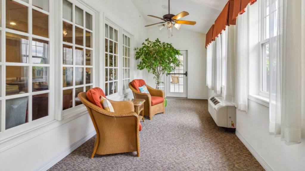 A bright sunroom with large windows, two wicker chairs with orange cushions, a small side table, a potted plant, ceiling fan, and neutral carpet flooring. White curtains and an air conditioning unit are also visible.