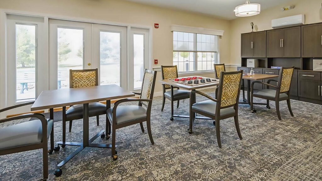 A bright, carpeted activity room with several tables and chairs, including a table set up for a board game, large windows, glass doors, and dark wood cabinets along one wall.