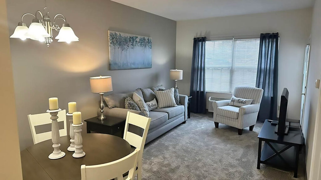 A cozy living room with a gray sofa, striped armchair, TV, and large window with blue curtains. A round dining table with candles and white chairs sits in the foreground under a chandelier. Neutral tones throughout.