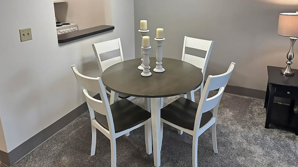 A small dining area with a round dark table, four white chairs, and two candle holders with three pillar candles. The room has grey carpet, light walls, and a lamp on a side table in the corner.