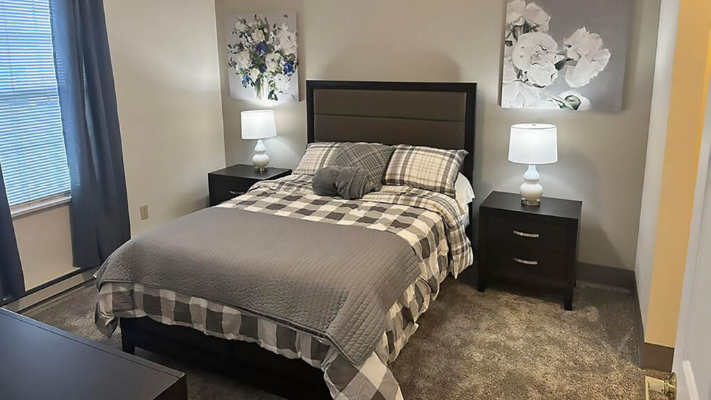 A cozy bedroom with a double bed covered in gray plaid bedding, two dark nightstands with white lamps, floral wall art above the bed, and a window with blue curtains letting in natural light.