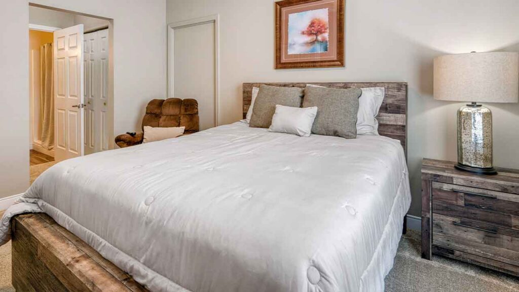 A bedroom with a queen-sized bed featuring a wood headboard and white bedding, next to a matching wood nightstand with a lamp and a brown armchair.