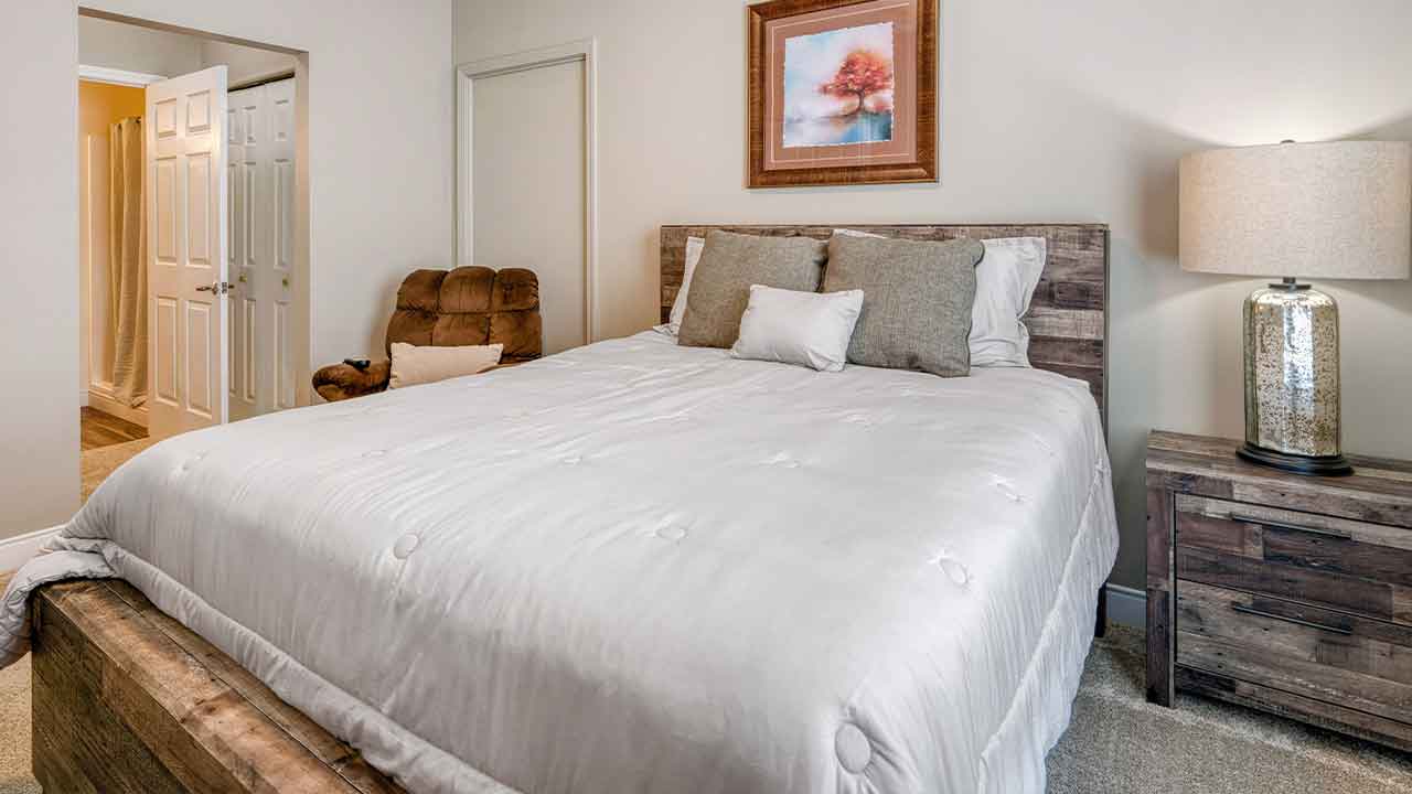 A bedroom with a queen-sized bed featuring a wood headboard and white bedding, next to a matching wood nightstand with a lamp and a brown armchair.