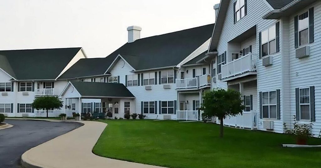 A two-story white apartment complex with green roofs, balconies, and large windows, surrounded by well-manicured grass and small trees alongside a curved paved walkway.