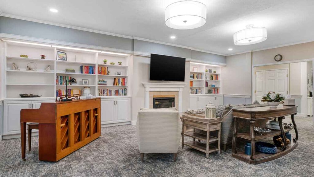 A living room featuring a wall of built-in white bookshelves, a central television above a fireplace, a wood-finish upright piano, and comfortable seating arranged on gray patterned carpet.