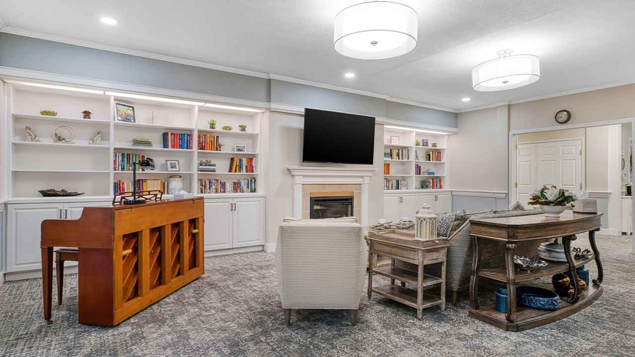 A living room featuring a wall of built-in white bookshelves, a central television above a fireplace, a wood-finish upright piano, and comfortable seating arranged on gray patterned carpet.