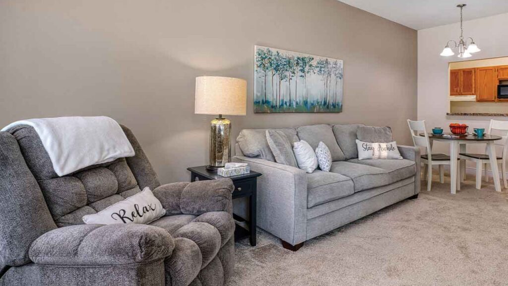 Living room with a gray reclining chair and sofa, both featuring throw pillows with text, next to a dining area.