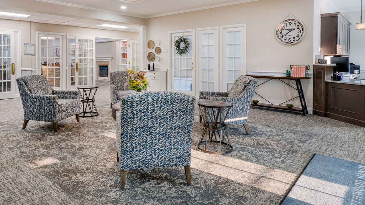 A bright, furnished lobby waiting area with several patterned armchairs, small side tables, a wall-mounted clock, and glass-paned double doors.