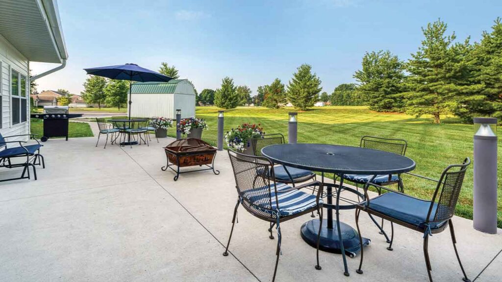 A concrete patio with two dining sets, a metal fire pit, and a grill, overlooking a large green lawn with trees.