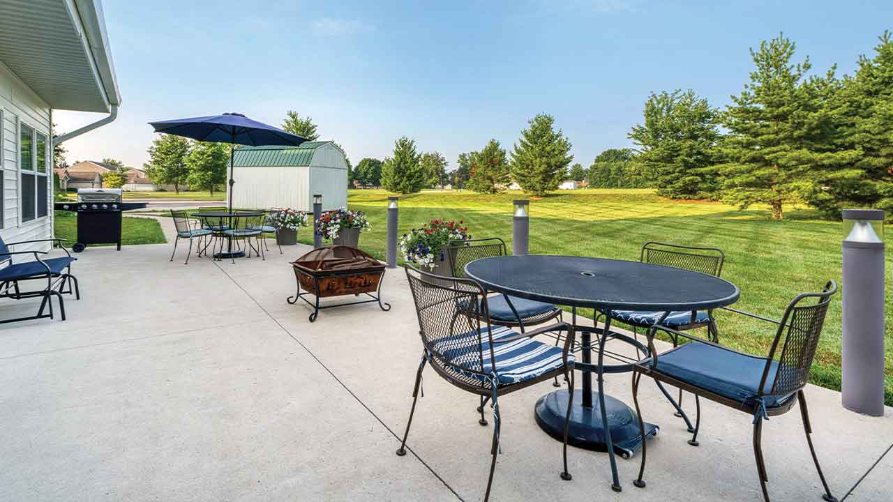 A concrete patio with two dining sets, a metal fire pit, and a grill, overlooking a large green lawn with trees.