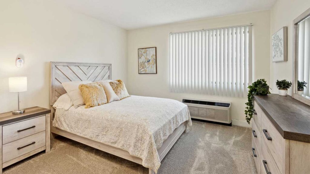 A bright, modern bedroom with a wooden bed, beige bedding, two pillows, a nightstand with a lamp, a dresser with plants, and a large window with vertical blinds letting in natural light.