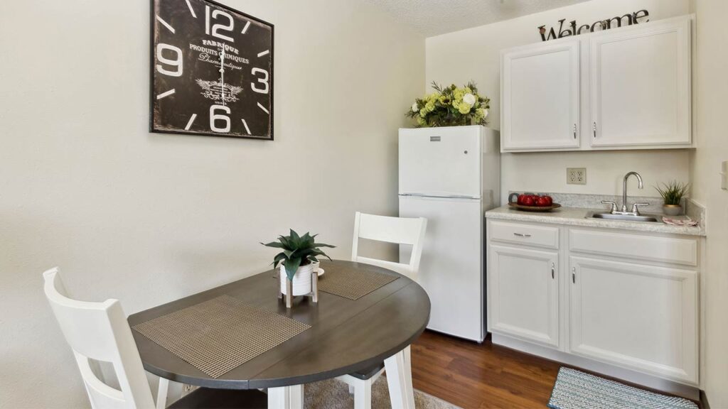 A small kitchen area with white cabinets, a white fridge, a round dark wood table set for two, white chairs, a large wall clock, and decorative plants on the counter and table.