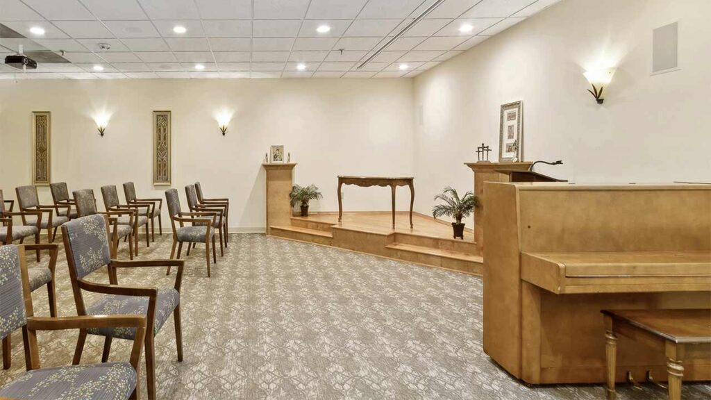 A small chapel with rows of chairs facing a raised wooden platform, a lectern, a piano, framed pictures on the wall, and light fixtures creating a calm and serene atmosphere.