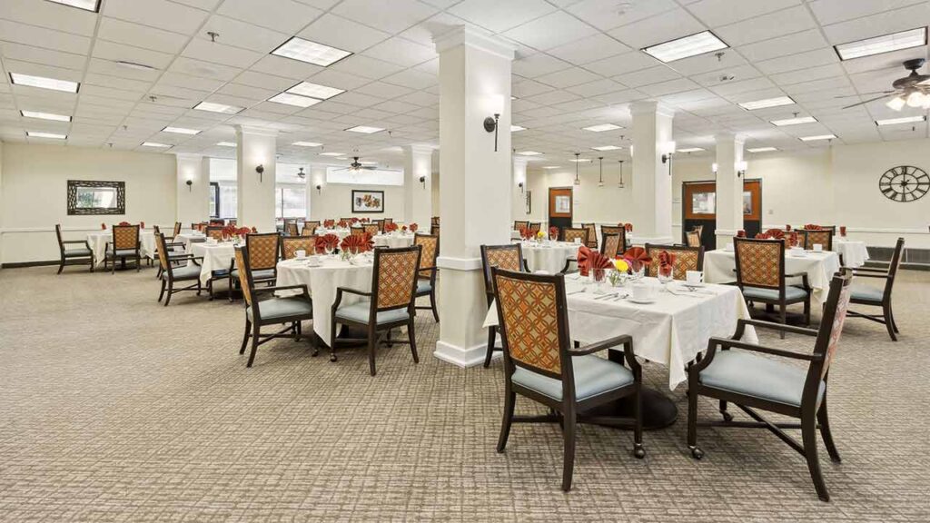 Spacious, well-lit dining room with neatly set tables covered in white tablecloths, surrounded by cushioned chairs. The room features columns, carpeted floors, and decorative wall art. No people are present.