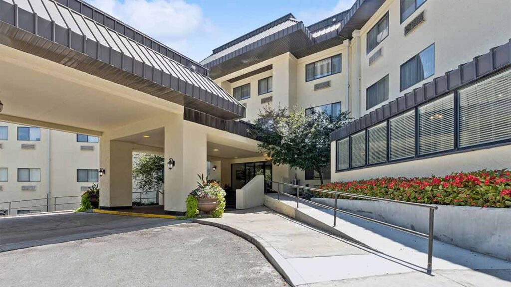 Accessible entrance of a modern building with a covered driveway, potted plants, a wheelchair ramp with railing, and a flower bed along the right side under large windows.