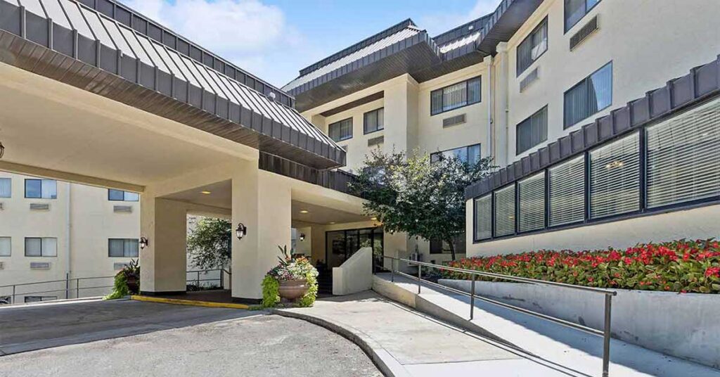 Entrance of a modern beige hotel with large windows, a covered driveway, and a ramp. Flowerbeds with red flowers line the right side, and a potted plant is near the entrance.