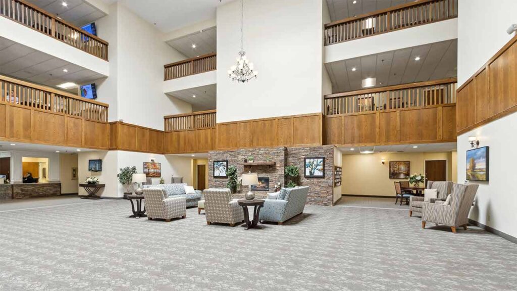 Spacious, well-lit lobby with high ceilings, a chandelier, and wood accents. Comfortable seating areas with armchairs and sofas are arranged on a patterned carpet. Reception desk and balconies are visible.