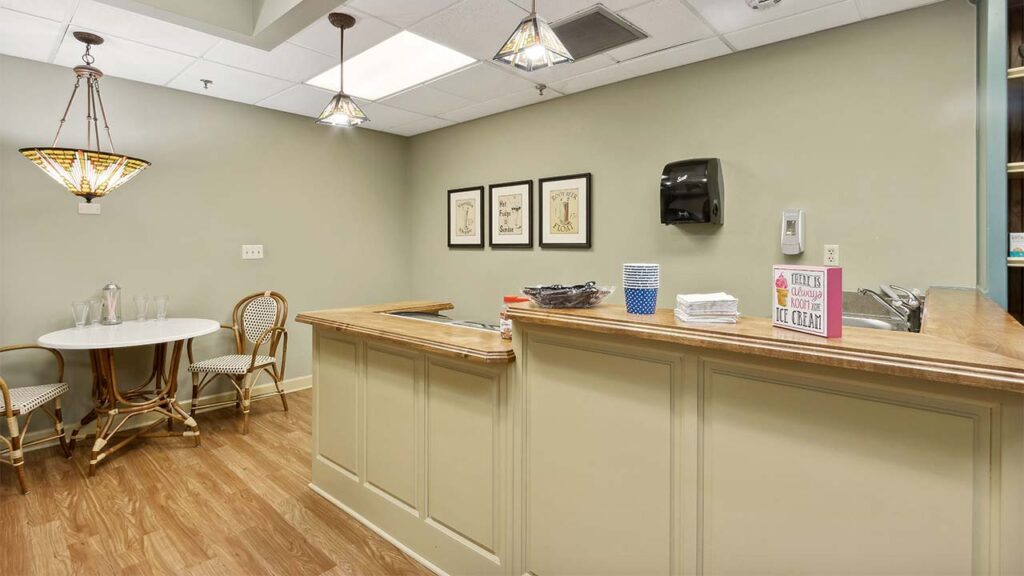 A small kitchen area with a wooden counter, two chairs, a round table, framed art on the wall, cups and a book on the counter, and light fixtures hanging from the ceiling. The floor is wood and walls are light green.