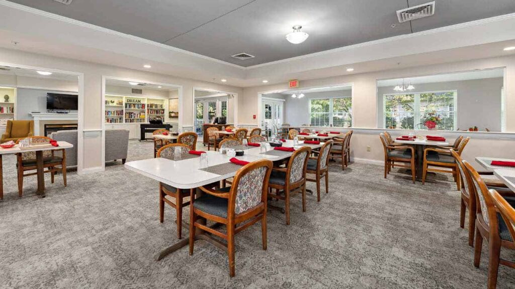 A spacious dining room with several wooden tables and chairs, each table set with red napkins. Large windows let in natural light, and bookshelves are visible in the background. The space appears clean and inviting.