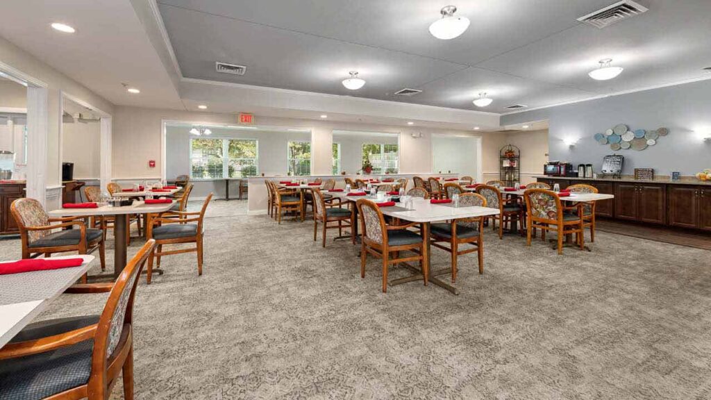 A spacious, well-lit dining room with several wooden tables and chairs, red napkins on tables, large windows, and a buffet area along the back wall. The room has neutral colors and bright ceiling lights.