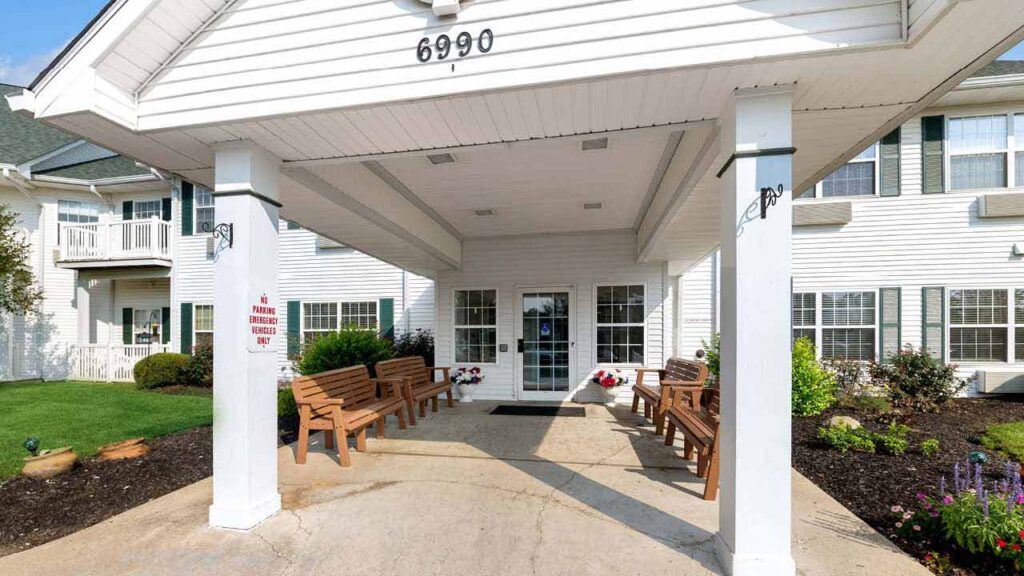 Covered entrance of a white building with benches on both sides, double glass doors, the number 6990 above the entrance, flowers, and neatly landscaped gardens.