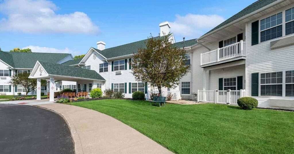 A large, two-story white building with green roofs, surrounded by well-kept lawns, shrubs, and a small tree. There is a covered entrance with chairs and a paved driveway curving in front of the building.