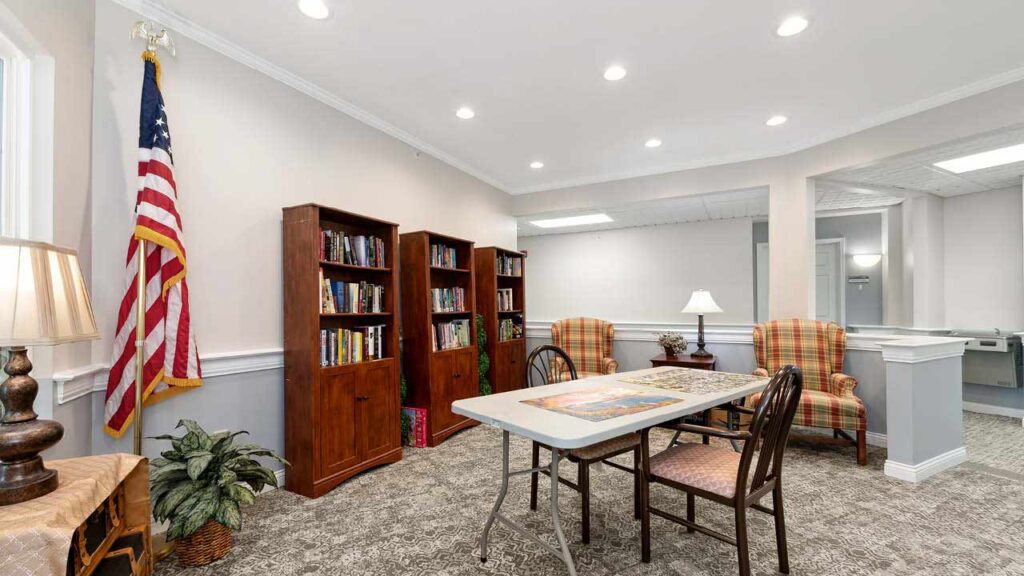 A cozy room with bookshelves, two plaid armchairs, a table with a puzzle in progress, wooden chairs, an American flag, a lamp, and a potted plant, all under bright ceiling lights.