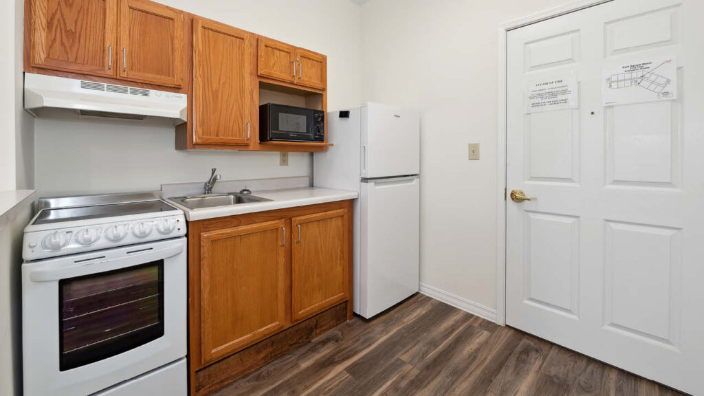 A small kitchen with wood cabinets, a white oven, a white refrigerator, a microwave, a stainless steel sink, and a white door with notices. The floor is wood-patterned laminate.