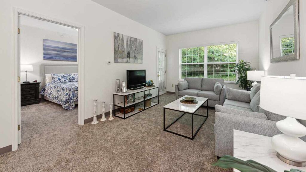 A bright living room with gray sofas, glass coffee table, TV on a shelf, and a bedroom visible through an open door. Large windows let in natural light, and green plants add a touch of color to the neutral decor.