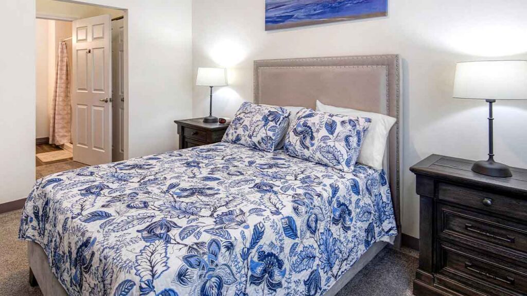 A bed with a blue and white floral patterned comforter and pillows, positioned between two dark wooden nightstands with lamps, in a room with an open doorway to a bathroom.