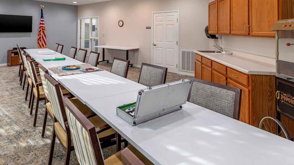 A long conference table with chairs in a community room featuring a kitchenette, a popcorn machine, and an American flag.