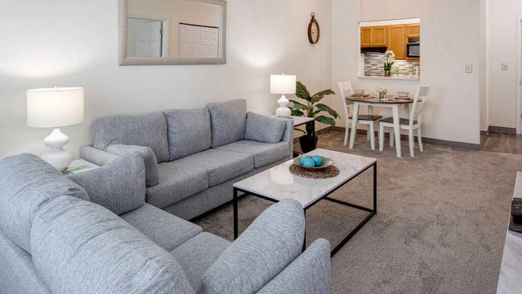 A living room with a gray sectional sofa, a marble-topped coffee table, and a small dining area in the background.
