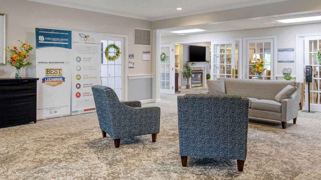 A lobby area featuring two patterned armchairs facing a gray sofa, with two informational banners standing to the left near glass doors.