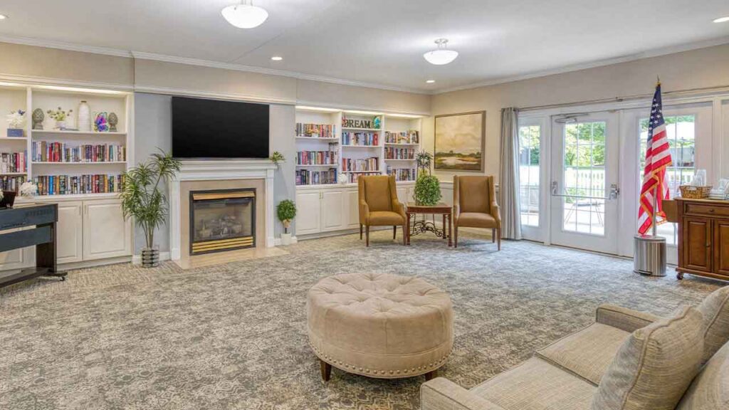 A spacious community lounge featuring a fireplace with a television above it, built-in bookshelves, two armchairs, a central round ottoman, and an American flag standing by glass doors.