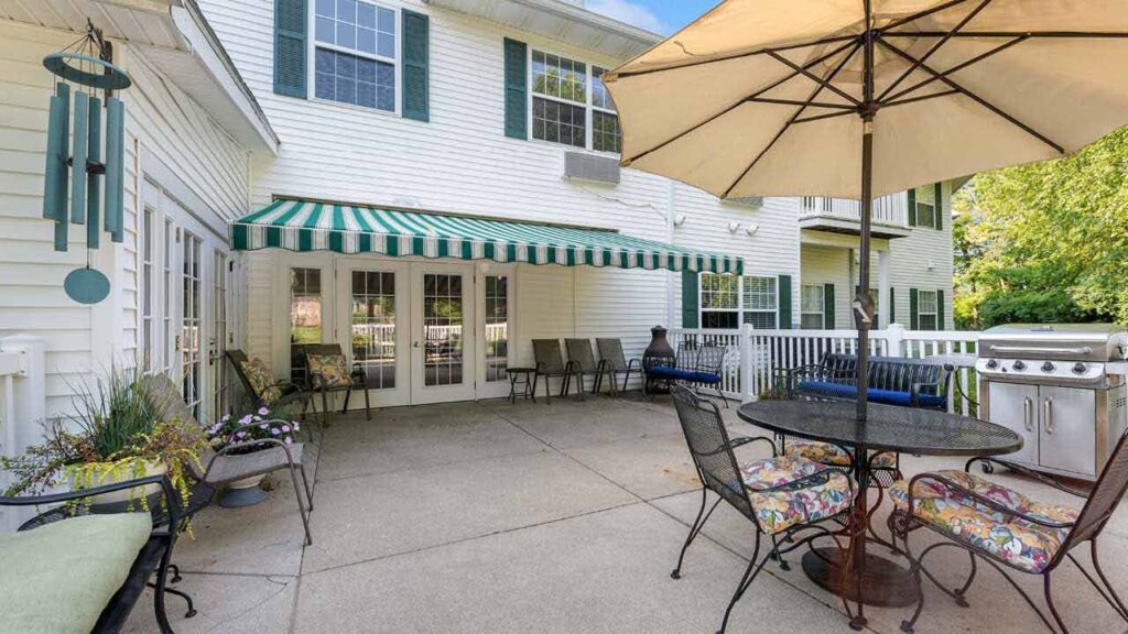 A spacious patio with a striped green awning, round table and chairs with colorful cushions, a grill, benches, potted plants, and a large umbrella outside a white two-story house with large windows.