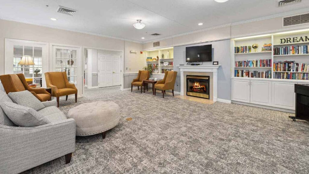 A cozy living room with armchairs, a sofa, and a round ottoman. Built-in bookshelves, a fireplace with a TV above it, and soft lighting create a warm, inviting atmosphere.