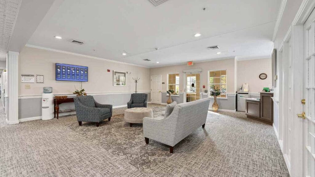 A spacious, well-lit lobby with two armchairs, a sofa, and a round ottoman arranged around a table. Large windows, double doors, and neutral decor create a welcoming atmosphere.