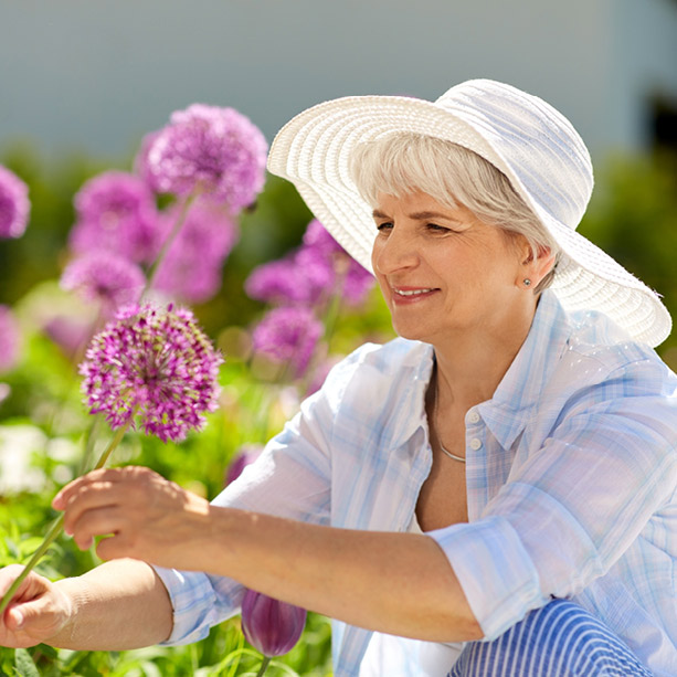 Older woman wearing a white sun hat and light blue shirt, smiling while tending to purple allium flowers in a garden on a sunny day.