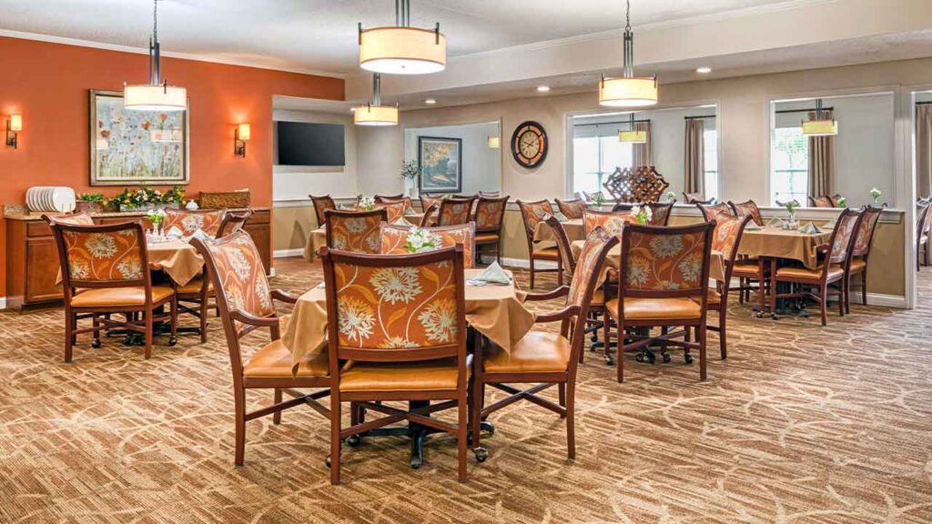 Dining room with several tables set with tan tablecloths and orange patterned chairs on a carpeted floor.