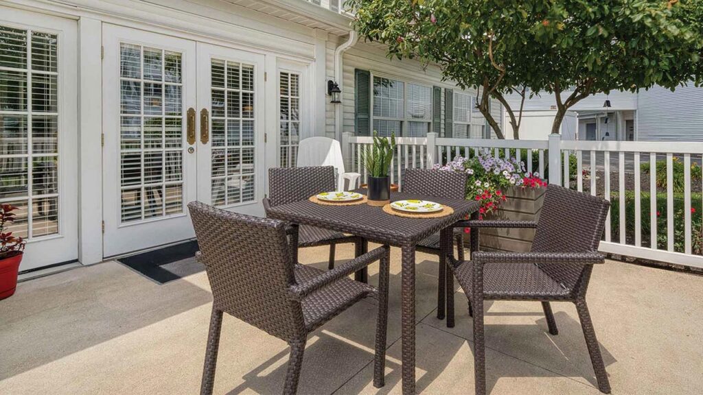 A patio set with a square table and four chairs on a concrete terrace in front of a house.