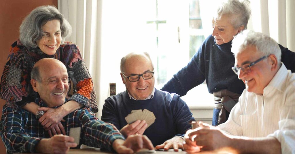 Five smiling older adults, three sitting at a table playing cards while two stand behind them, chatting and enjoying each other's company in a warmly lit room.