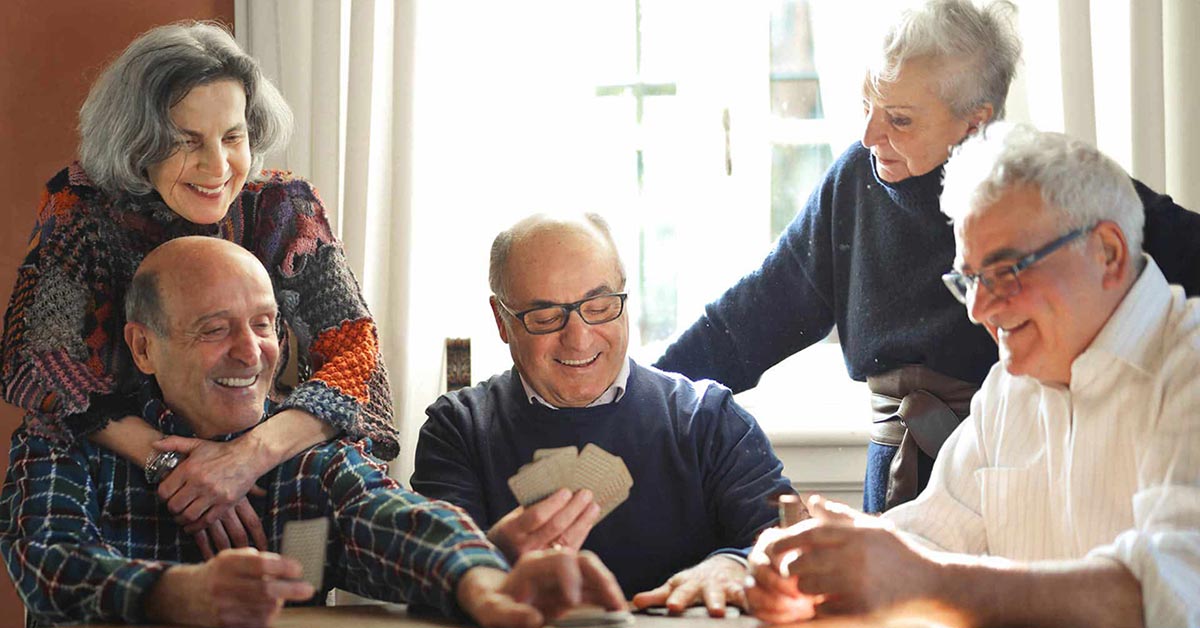 Five smiling older adults, three sitting at a table playing cards while two stand behind them, chatting and enjoying each other's company in a warmly lit room.