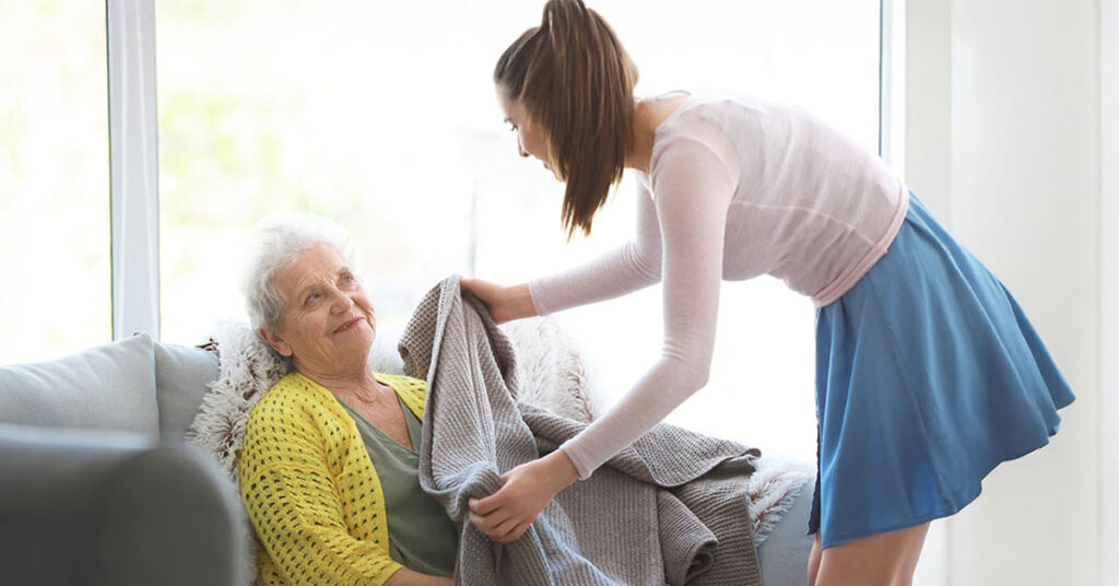 A young woman in a blue skirt gently covers an elderly woman sitting on a couch with a gray blanket. The elderly woman, wearing a yellow sweater, looks up at the young woman and smiles. Bright light comes through the window behind them.