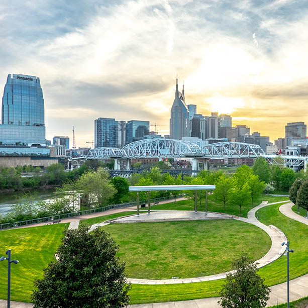 A cityscape view of Nashville, Tennessee, featuring a park with green lawns, a white bridge over a river, modern skyscrapers, and the iconic AT&T building against a bright, partly cloudy sky at sunset.