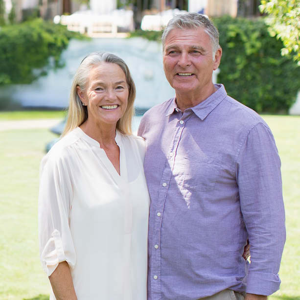 An older couple stands together outdoors, smiling at the camera. The woman wears a white blouse and the man wears a light purple shirt. Lush greenery and a blurred building are visible in the background.
